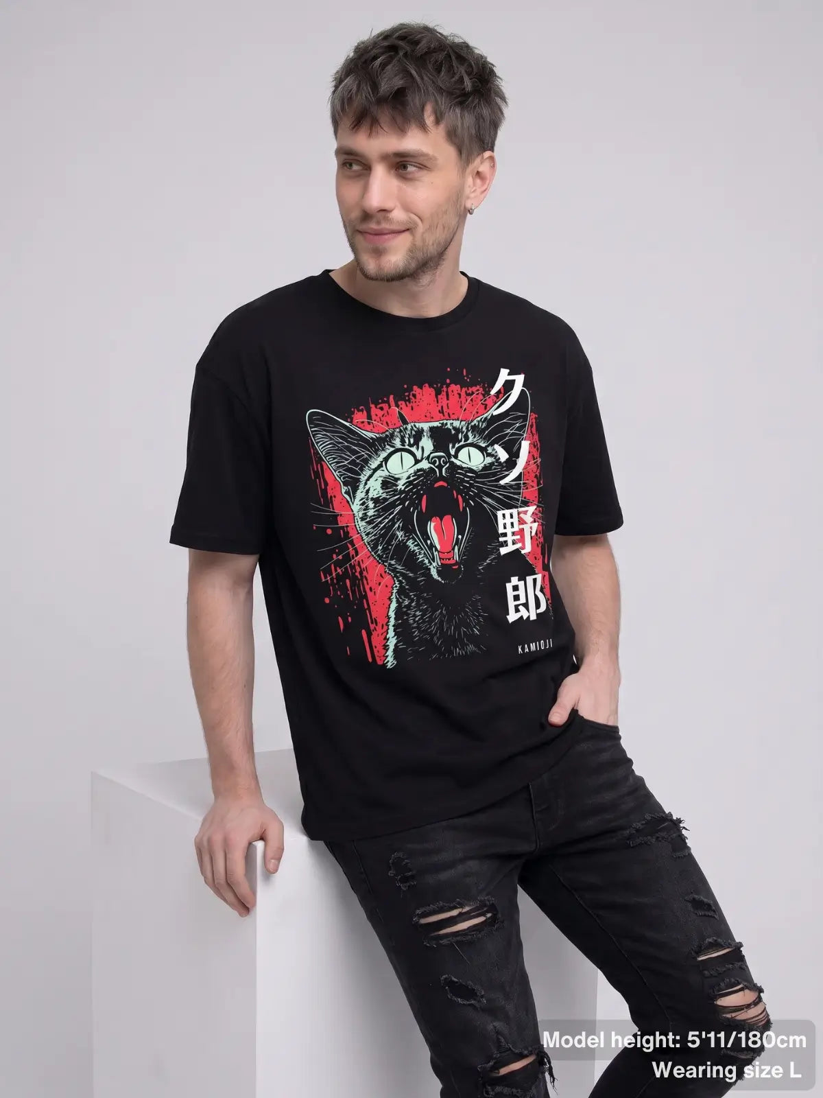 A man wearing a black graphic t-shirt with a stylized cat illustration in red, white, and green, paired with distressed black jeans, leaning against a white block in a studio setting.